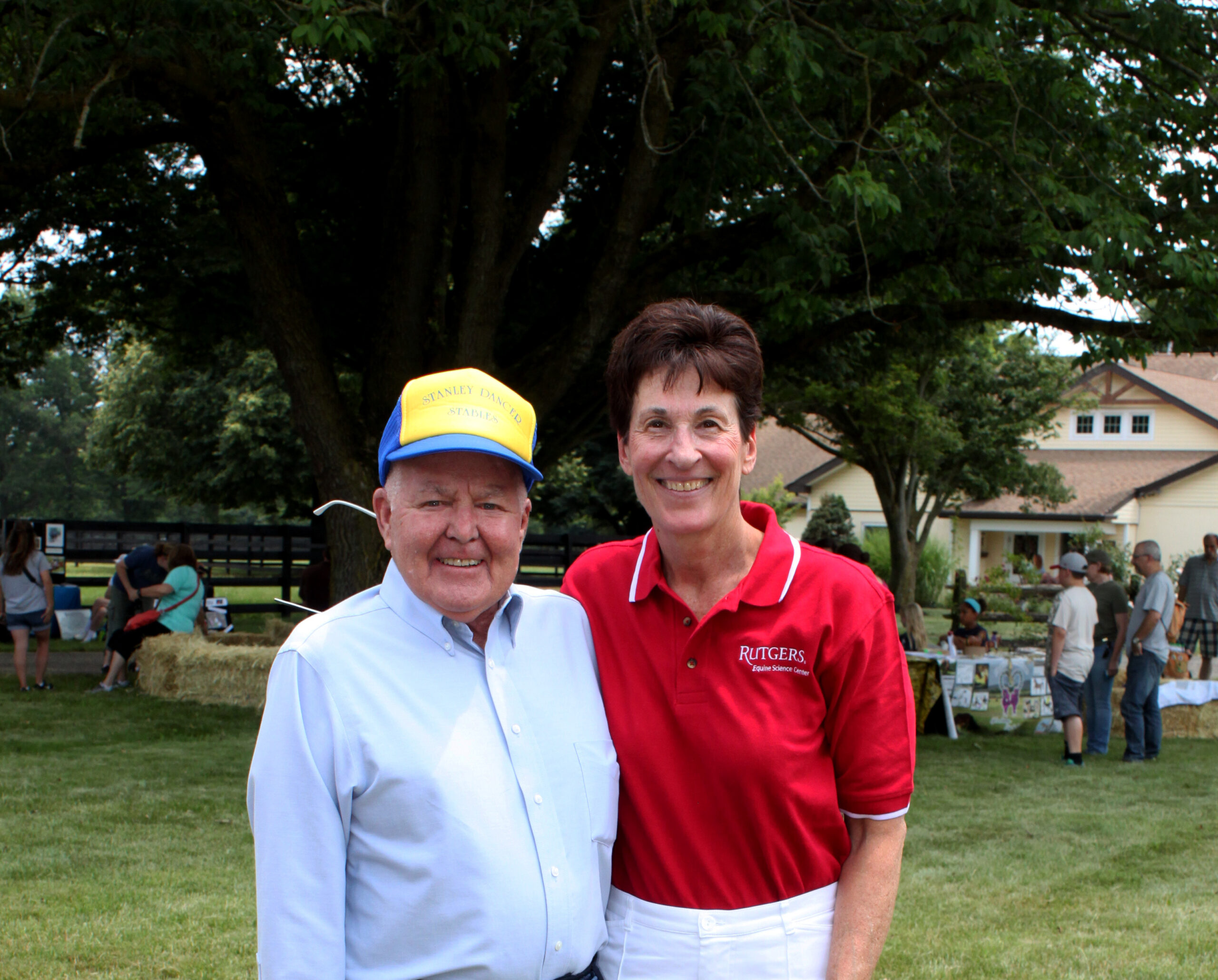 Two people smiling together outdoors, with one wearing a blue and yellow cap and the other in a red Rutgers shirt, surrounded by people and greenery in the background.