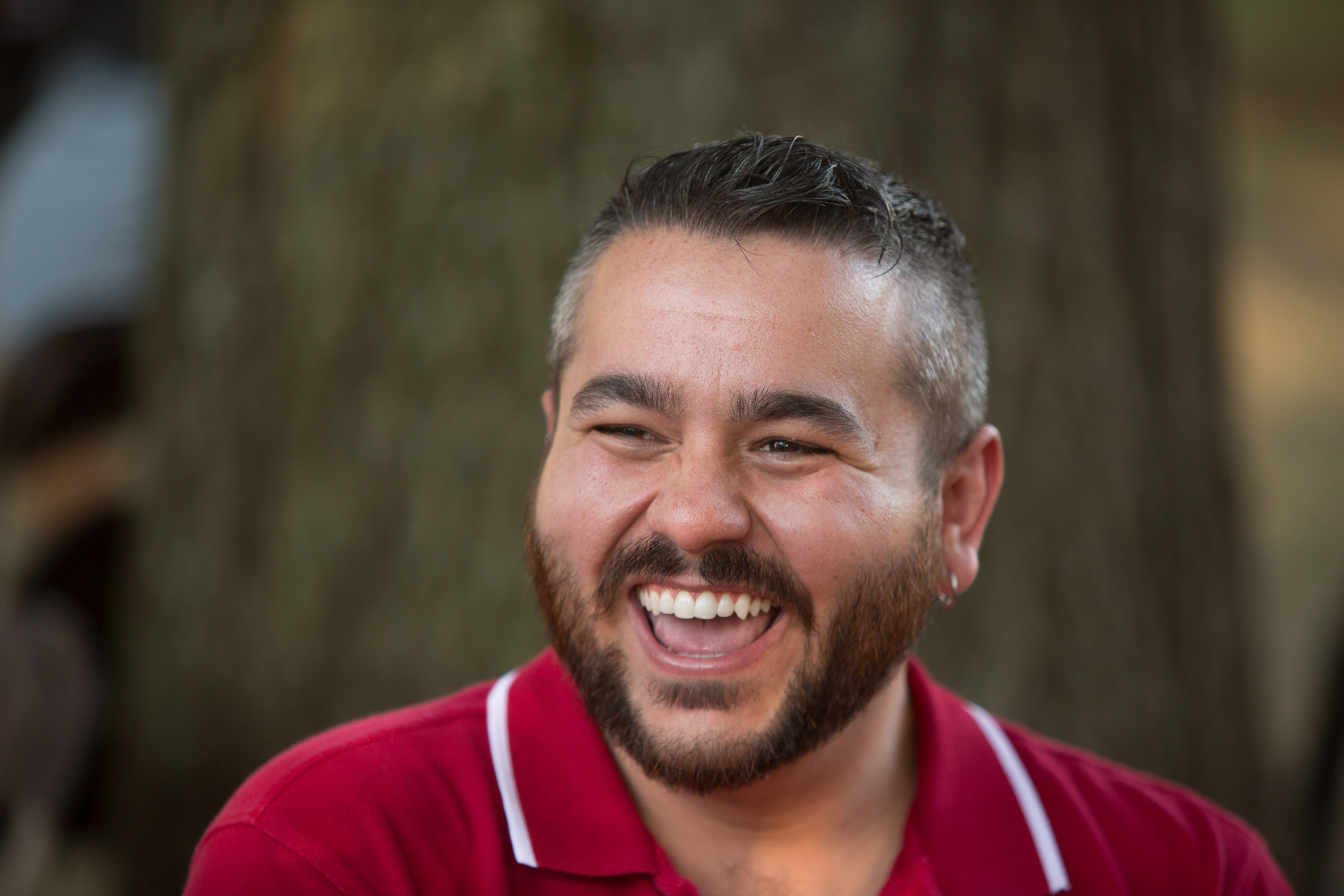 A man with a short beard and earrings laughing joyfully while wearing a red shirt, sitting outdoors against a blurred natural background.